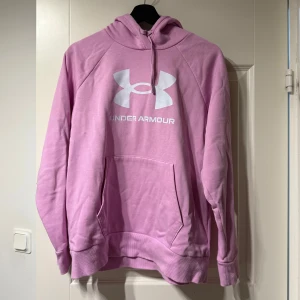 Rosa hoodie från Under Armour - Säljer en rosa hoodie från Under Armour med stor logga på bröstet. Tröjan har en klassisk känguruficka och justerbar huva. Perfekt för en avslappnad stil.