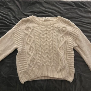 Beige stickad tröja från KappAhl - Mysig beige stickad tröja från KappAhl med kabelstickat mönster. Perfekt för kyliga dagar med sin varma och bekväma design. Tröjan har långa ärmar och ribbade kanter.
