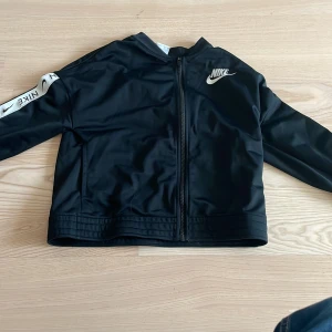 Svart hodie från Nike - Säljer en svart hodie från Nike med dragkedja och logga på bröstet. Hodienhar långa ärmar och en stilren design. Perfekt för en sportig look.