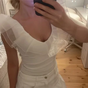 Vit topp med volangdetaljer - Säljer en elegant vit topp med volangdetaljer vid axlarna. Toppen har en djup v-ringning och är figurnära, vilket ger en smickrande passform. Perfekt för en stilren look. Helt oanvänd body med prislapp kvar 