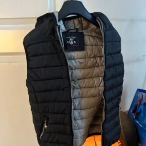 Svart dunväst från O&S - Säljer en svart dunväst från O&S med huva och dragkedja. Västen har en quiltad design och praktiska fickor med dragkedjor. Perfekt för kyligare dagar. Står storlek L men är mer som en S/XS. 