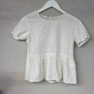 Vit spetsblus från Sinsay - Säljer en söt vit spetsblus från Sinsay i storlek XS. Blusen har korta ärmar och ett fint blommigt mönster. Den har en volangdetalj vid midjan som ger en feminin touch. Perfekt för en somrig look! 