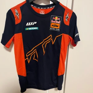 Svart och orange t-shirt från Red Bull KTM Racing Team - Snygg svart och orange t-shirt från Red Bull KTM Racing Team med tryck av logotyper på fram- och baksidan. Perfekt för racingfans som vill visa sitt stöd. Kortärmad design med en sportig look.