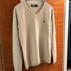 Beige tröja från Polo Ralph Lauren - Säljer en stilren beige tröja från Polo Ralph Lauren med v-ringning och det klassiska logotypbroderiet på bröstet. Tröjan är långärmad och har ribbade kanter vid ärmslut och nederkant. Perfekt för en avslappnad men elegant look.