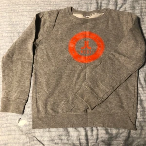 Grå sweatshirt med orange tryck - Säljer en grå sweatshirt med ett coolt orange tryck på bröstet. Tröjan har långa ärmar och är perfekt för en avslappnad stil. Den är mjuk och bekväm, idealisk för vardagsbruk.