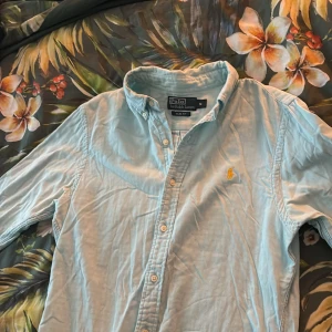 Ljusblå skjorta från Ralph Lauren - Mitt i flytt så tyvär skrynklig bilder. Snygg ljusblå skjorta från Ralph Lauren i slim fit. Skjortan har långa ärmar och knappar framtill. Den är broderad med den klassiska loggan i gult på bröstet. Perfekt för en stilren look!
