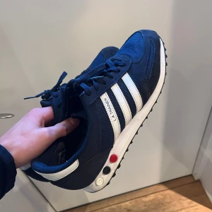 Blå Adidas LA Trainer sneakers - Snygga blå Adidas LA Trainer sneakers med vita ränder och snörning. Skorna har en klassisk design med en robust sula och detaljer i rött och vitt på sidan. Perfekta för en sportig look.