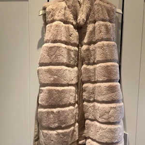 Beige pälsväst från Amisu - Säljer en lyxig beige pälsväst från Amisu. Västen har en elegant design med ett bälte i midjan för en snygg passform. Perfekt för att hålla sig varm och stilren under kyligare dagar.