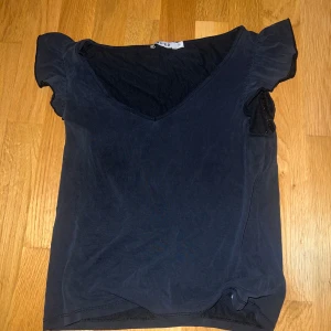 Svart topp från NA-KD - Säljer en stilren svart topp från NA-KD i storlek XS. Toppen har en v-ringad design och korta ärmar med en liten volangdetalj. Perfekt för en enkel och snygg look. 