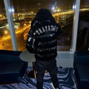 Svart pufferjacka från Trapstar - Säljer en svart pufferjacka från Trapstar med glansig yta och stor logga på ryggen. Jackan har dragkedja framtill och är perfekt för kyliga dagar. Materialet är en blandning av PU och polyester vilket ger en skön och varm känsla.