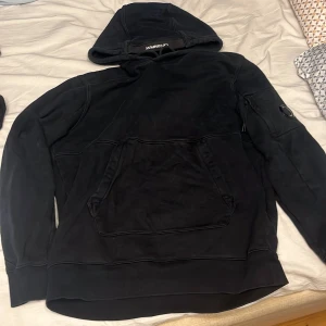 Svart hoodie från C.P. Company - Säljer en svart hoodie från C.P. Company med en praktisk magficka och huva. Den har en unik detalj med en rund logotyp på ärmen. Perfekt för en stilren och bekväm look. Storlek M