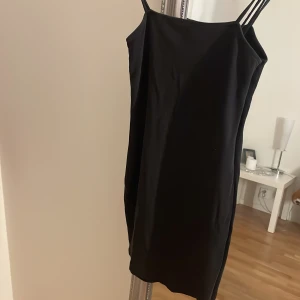 Svart axelbandslös klänning - Snygg svart klänning med smala axelband och en elegant, enkel design. Perfekt för en stilren look. Klänningen har en figurnära passform och är knälång.