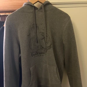 Grå hoodie med tryck - Säljer en grå hoodie med ett marint tryck av en fyr och ett ankare på framsidan. Tröjan har en klassisk känguruficka och justerbar huva med dragsko. Perfekt för en avslappnad stil.