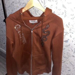 Brun hoodie med ormmotiv från Trapper - Säljer en brun hoodie från Trapper med dragkedja och en cool ormmotiv i paljetter på framsidan. Hoodien har långa ärmar och snörning vid huvan. Perfekt för en avslappnad stil.