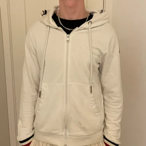 Vit hoodie från Moncler - Säljer en stilren vit hoodie från Moncler med dragkedja och justerbar huva. Tröjan har Moncler-logga på ärmen och ribbade muddar. Perfekt för en avslappnad look. Den är använd ett par gånger och de finns en liten fläck på framsidan men den går att tvätta bort! De finns även två små små hål längst ner på armarna men inget man lägger märke till och ingen större skada! Skicka dm vid intresse! Pris kan diskuteras vid snabb affär!
