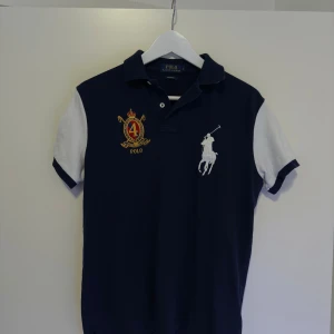 Mörkblå pikétröja från Ralph Lauren - Säljer en snygg mörkblå pikétröja från Ralph Lauren med vita ärmar och ett stort broderat logotypmotiv på bröstet. Tröjan har en klassisk krage och knappar framtill. Perfekt för en stilren look.