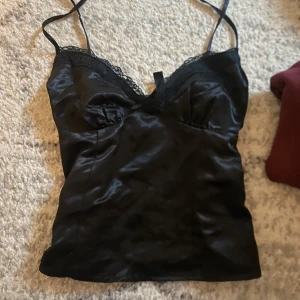 Svart satinlinne med spetsdetaljer - Snyggt svart satinlinne med smala axelband och spetsdetaljer vid urringningen. Perfekt för en stilren och elegant look.