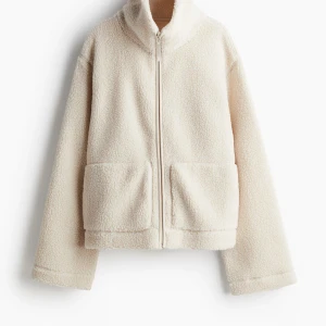 Beige teddyjacka med dragkedja - Mysig beige teddyjacka med dragkedja framtill och stora fickor. Perfekt för kyliga dagar med sin fluffiga och varma design. Jackan har en hög krage och långa ärmar för extra värme.