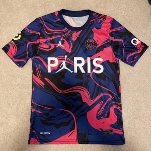 Paris Saint-Germain fotbollströja - Snygg PSG-tröja i samarbete med Jordan. Den har ett unikt mönster i blått, rosa och svart med vit text.