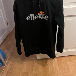 Svart hoodie från Ellesse - Säljer en svart hoodie från Ellesse med logotyp i vitt och orange på bröstet. Tröjan har långa ärmar och en praktisk ficka framtill. Perfekt för en avslappnad stil.