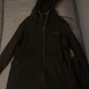 Svart hoodie från Hugo Boss - Säljer en stilren svart hoodie från Hugo Boss med dragkedja och huva. Perfekt för en avslappnad look. Den har långa ärmar och en diskret logga på bröstet. Passar bra till både jeans och joggers.