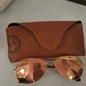 Solglasögon från Ray-Ban - Snygga solglasögon från Ray-Ban med guldiga bågar och spegelglas i rosa/rose nyans. Klassisk pilotmodell som passar perfekt för soliga dagar. Kommer med ett brunt fodral för säker förvaring.