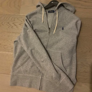 RAPLH LAUREN ZIP - Säljer en klassisk grå hoodie från Polo Ralph Lauren med dragkedja och huva. Den har långa ärmar och en liten broderad logga på bröstet. Perfekt för en avslappnad stil. Väldigt bra skick och inga defekter alls