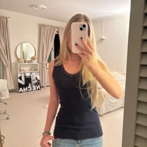 Svart ribbad linnetopp med spetsdetaljer - Snygg svart ribbad linnetopp med spetsdetaljer vid halsringningen. Perfekt för en stilren look och kan enkelt matchas med olika outfits. Toppen har en tight passform och ärmlös design.