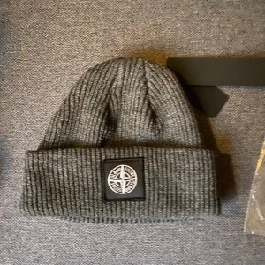 Grå  mössa från Stone Island - Snygg mössa från Stone Island med ett broderat märke framtill. Perfekt för kyliga dagar och ger en stilren look. Mössan är tillverkad i ett mjukt och bekvämt material.