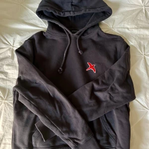 Svart hoodie från Axel Arigato - Säljer en stilren svart hoodie från Axel Arigato med en röd fågelbrodyr på bröstet. Tröjan har en klassisk design med huva och dragsko samt ribbade muddar vid ärmslut och nederkant. Perfekt för en avslappnad look.