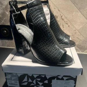Svarta klackskor från Tiamo - Snygga svarta klackskor från Tiamo med krokodilpräglat mönster. . Skorna har en bred klack och justerbart spänne runt ankeln. Perfekta för att ge en stilfull touch till din outfit. Sommarens Dansa-Hela-Natten-Lång klackar! Aldrig använda, endast provade.  Nypris 699:-  Se även min andra annons där jag säljer de i beige.