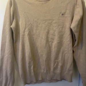 Beige tröja från GANT - Säljer en stilren beige tröja från GANT med rund halsringning och diskret logga på bröstet. Tröjan har långa ärmar och ribbade muddar vid ärmslut och nederkant. Perfekt för en avslappnad och snygg look.