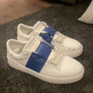 Vita sneakers med blå detalj - Snygga vita sneakers från Valentino med en iögonfallande blå detalj över snörningen. Skorna har en klassisk design med rund tå och platt sula, perfekt för en stilren look. Valentino påse och ett valentino kort ingår också.