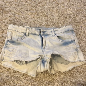 Ljusblå jeansshorts från Sisley - Snygga ljusblå jeansshorts från Sisley med broderade blommor framtill. De har en lågmidjad passform och fransiga kanter för en avslappnad look. Perfekta för varma sommardagar!