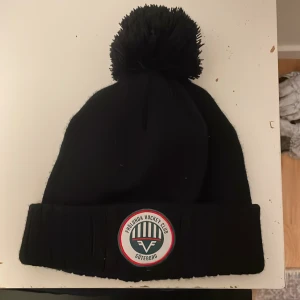 Svart mössa med Frölunda Hockey Club-logga - Snygg svart mössa med stor tofs på toppen och Frölunda Hockey Club-logga framtill. Perfekt för att hålla värmen under kalla dagar och visa ditt stöd för laget.