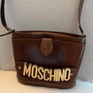 Brun axelväska från Moschino - 100% äkta Moschino vintageväska - vintage bag. Köptes för ca 15 år sedan som vintage så kvitto finns ej men 100% äkta.  Moschino i gulddetaljer. Rödbrunt läder. Lång axelrem. Ytterficka som stängs med knapp och liten innerficka som stängs med dragkedja.  Mått ca: 25cm x 17cm.  Hämtas eller postas med spårbarhet.