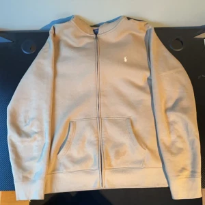 Ralph Lauren Zip Up Hoodie - Säljer en stilren beige hoodie från Polo Ralph Lauren med dragkedja och det klassiska broderade logotypen på bröstet. Tröjan har en bekväm passform och praktiska fickor framtill. Perfekt för en avslappnad look. 