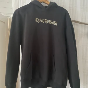 Svart Iron Maiden hoodie från H&M - Säljer en svart hoodie från H&M med Iron Maiden-tryck. Framsidan har bandets logga och baksidan pryds av ett stort, detaljerat motiv i blått och vitt. Perfekt för fans av bandet! Hoodien har en avslappnad passform och en klassisk känguruficka framtill. Storleken är S i man storlek så den är större