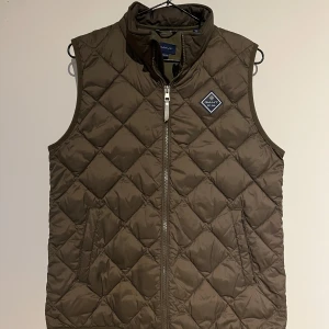 GANT väst - Snygg oliv grön väst från GANT med dragkedja. Perfekt för lager-på-lager under kyligare dagar. Västen har en diskret logotyp på bröstet och är lätt att matcha med olika outfits. Priset kan diskuteras.