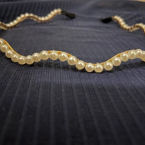 Pannband med pärlor - Elegant pannband dekorerat med vita pärlor på en gyllene bas. Perfekt för att ge en stilfull touch till din look. Justerbart med svarta band i ändarna. Inte använd