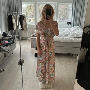 Blommig långklänning från Mix By - Säljer en romantisk långklänning från Mix By i en härlig rosa nyans med blommigt mönster. Klänningen har en v-ringad design och knytning i midjan som ger en smickrande passform. Perfekt för sommarens festligheter!