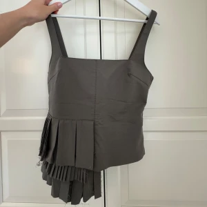 Brun topp med plisserad detalj - Säljer denna militärgröna toppen från Zara. Toppen är i nyskick och har bara använts en gång. Superfin för både fest och vardag! Finns en dragkedja på sidan.