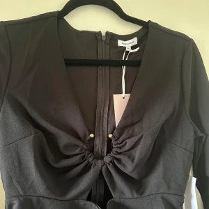 Svart klänning från House of CB - Säljer en elegant svart klänning från Bubbleroom. Den har en snygg v-ringning med knytning framtill och långa ärmar. Perfekt för festliga tillfällen! Ny med etiket på