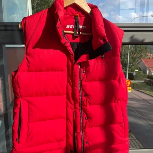 Röd dunväst från Everest - Säljer en snygg och varm röd dunväst från Everest. Perfekt för kyliga dagar när du vill hålla värmen utan att bära en jacka. Västen har en dragkedja och knappar framtill för extra skydd mot vinden. Den är i nyskick och redo för nya äventyr!