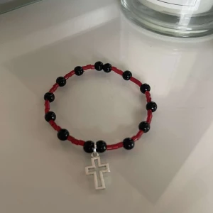 Armband med korsberlock - Säljer ett coolt armband med svarta och röda pärlor. Armbandet har en silverfärgad korsberlock som hänger i mitten. Perfekt för att ge en edgy touch till din outfit!
