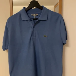 Blå pikétröja från Lacoste - Säljer en klassisk blå pikétröja från Lacoste med den ikoniska krokodilloggan på bröstet. Tröjan har korta ärmar och en traditionell krage med knappar. Perfekt för en stilren och avslappnad look.