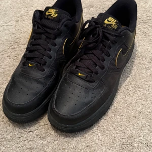 Svarta Nike Air Force 1 med gula detaljer - Snygga svarta Nike Air Force 1 med gula detaljer. Skorna har en klassisk design med snörning och är tillverkade i skinn. Perfekta för en stilren look.