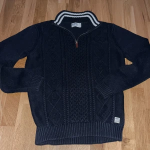 Mörkblå stickad tröja från Dobber - Säljer en snygg mörkblå stickad tröja från Dobber med dragkedja och ribbad krage i vitt och blått. Tröjan har ett stilrent mönster och långa ärmar, perfekt för kyligare dagar. Passar bra till både jeans och chinos.