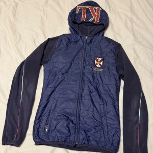 Blå jacka från Kingsland/ junior - Säljer en blå jacka från Kingsland med huva och dragkedja. Jackan har långa ärmar med röda och vita detaljer samt ett broderat emblem på bröstet. Baksidan har en vit panel med Kingsland-loggan i rött. Perfekt för en sportig look!