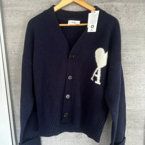 Ami Paris Cardigan - Snygg mörkblå kofta från Ami med ett vitt hjärta och bokstaven 'A' på bröstet. Koftan har en v-ringad design och knappar framtill. Perfekt för en stilren look.